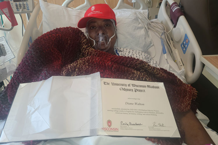 Diane lying in a hospital bed wearing a respirator, and holding her Odyssey graduate diploma