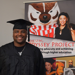 Anthony in his cap and gown posing next to Odyssey Project signage