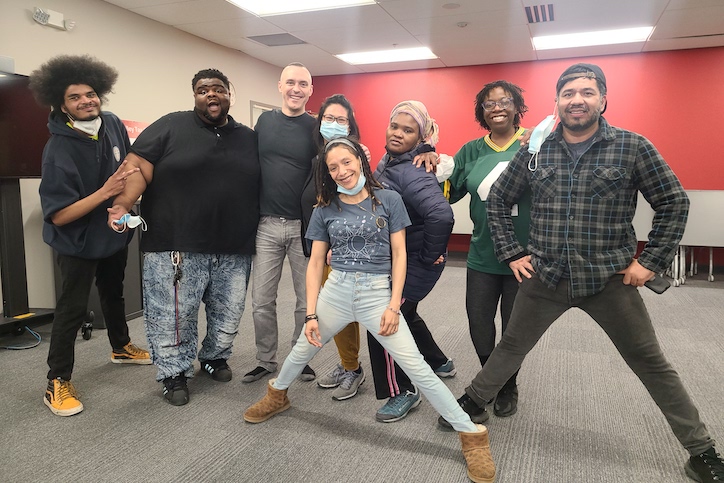 Eight adult Onward Odyssey students of varying ages and ethnicities smile while standing in a classroom