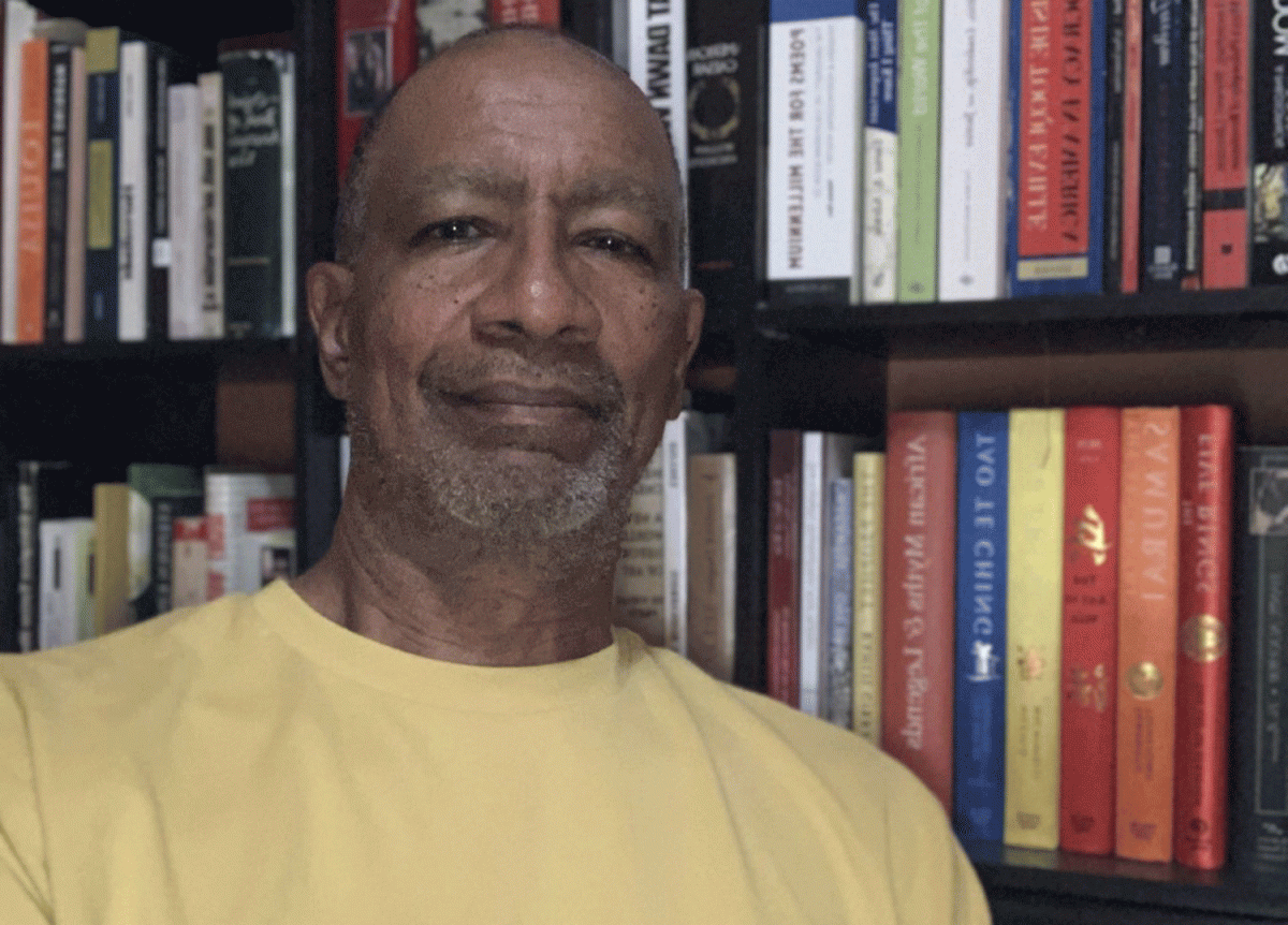 Robert Taliaferro standing, smiling, in front of his bookcase, wearing a yellow shirt