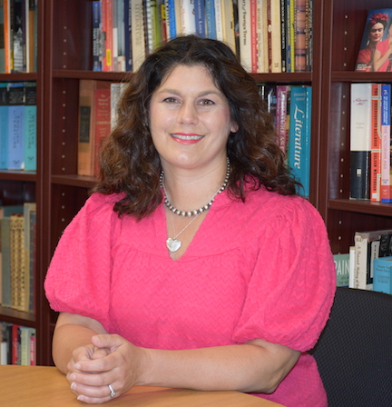 UW Odyssey instructor Erin Cellelo sits at a table smiling in front of bookshelf filled with books in background