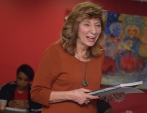 Jean Feraca stands smiling while teaching her class. She is holding a notebook in her hands. A student sits at a table in the background, looking down.
