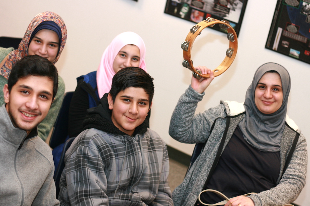 A seated woman holds up a tambourine. Two young women and two young men sit next to her. All of the women are wearing headscarves.