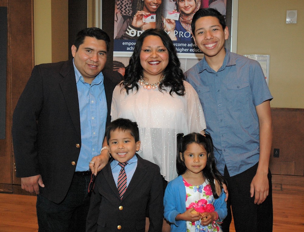 A woman stands smiling with her husband, young adult son, young son and young daughter. All are smiling for the camera.