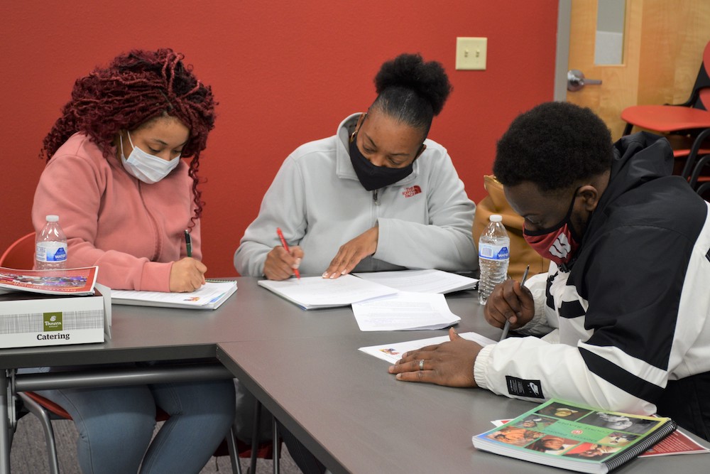 Three UW–Madison Odyssey Project students sit writing at tables. They are all wearing masks. 